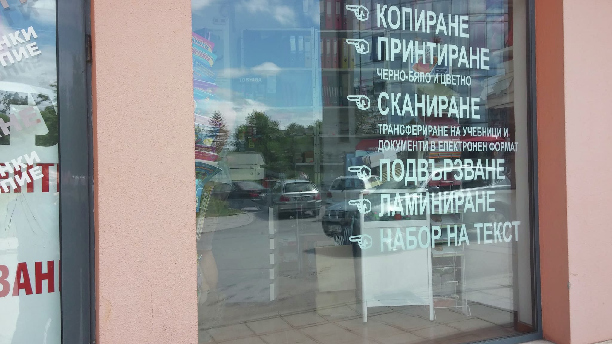 Витрина на магазин с рекламен надпис от фолио, показващ услуги като копиране, печат, сканиране, подвързване, ламиниране и набор на текст, с икони за посока.
