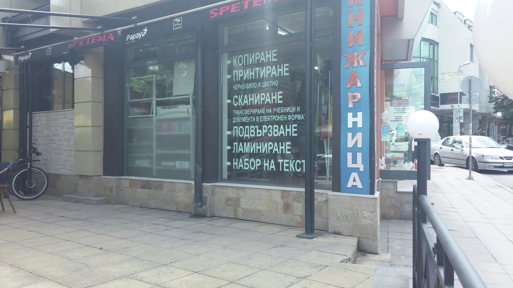 Витрина на магазин „Спетема“ с рекламен надпис от фолио, изброяващ печатни услуги като копиране, принтиране, сканиране и ламиниране.