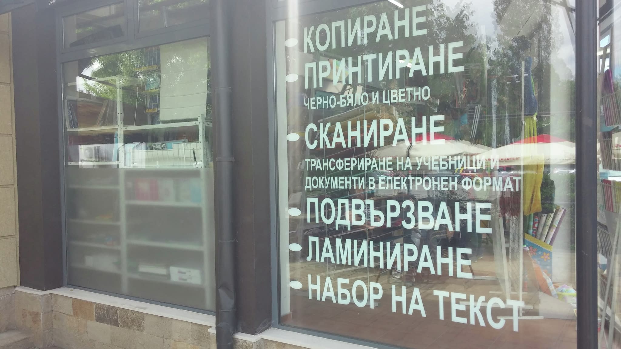 Рекламен надпис с фолио на витрина, показващ предлагани услуги като копиране, печат, сканиране, подвързване, ламиниране и набор на текст.