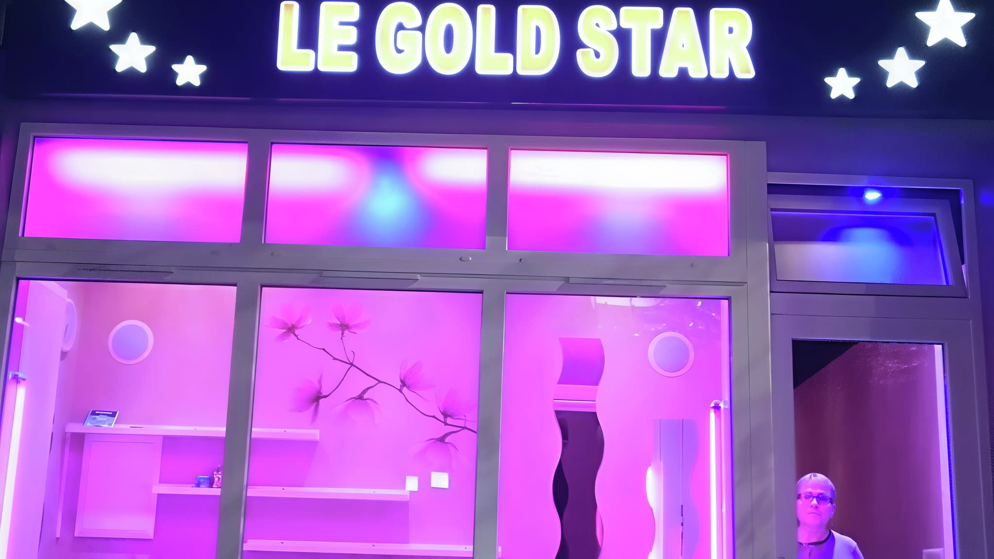 Осветена витрина на фризьорски салон „Le Gold Star“ с розов интериор, фризьорски столове и декоративно LED осветление.