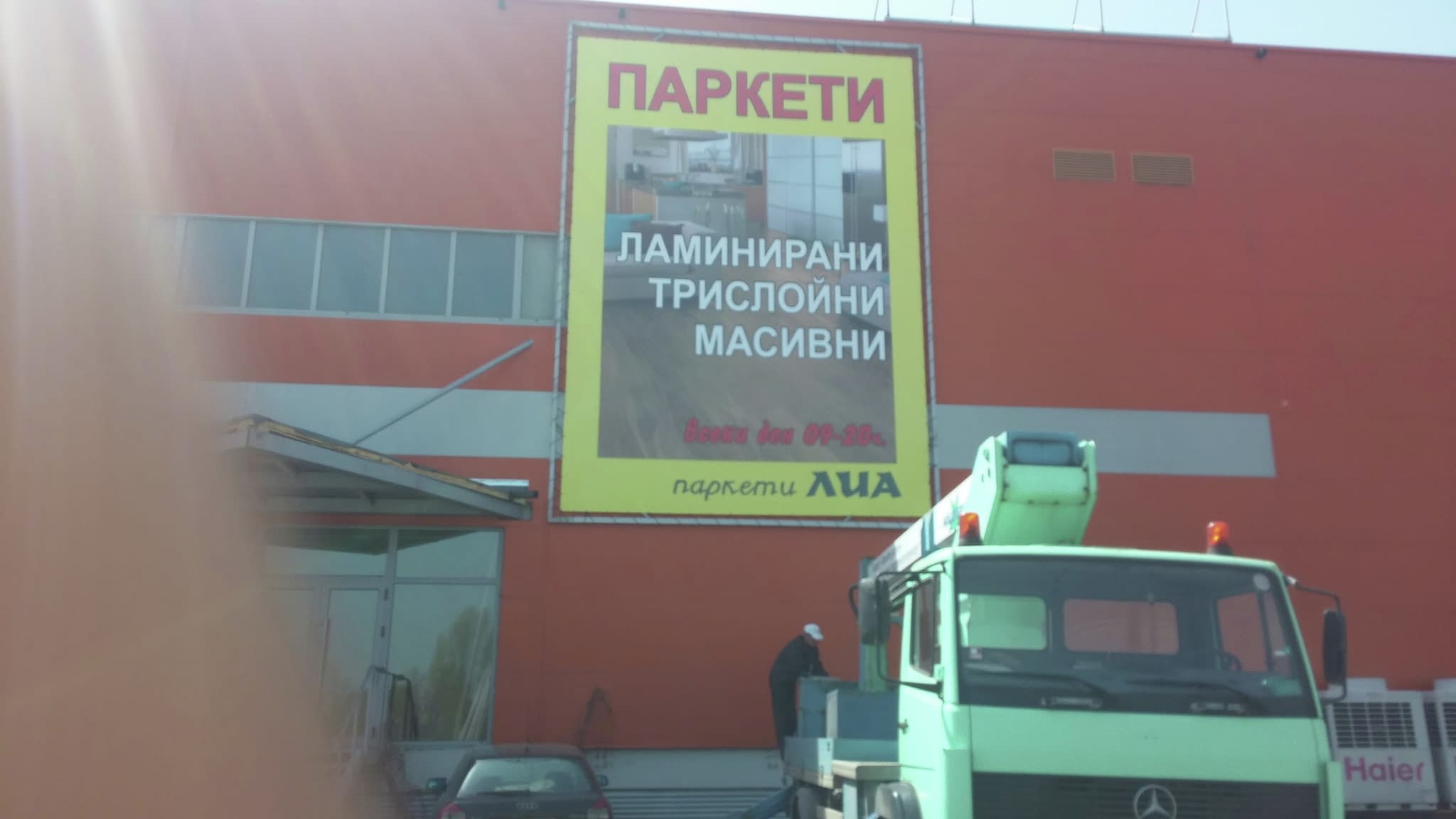 Голяма винилова реклама на паркети, поставена върху фасада на оранжева сграда, с паркиран зелен камион пред входа на магазина.