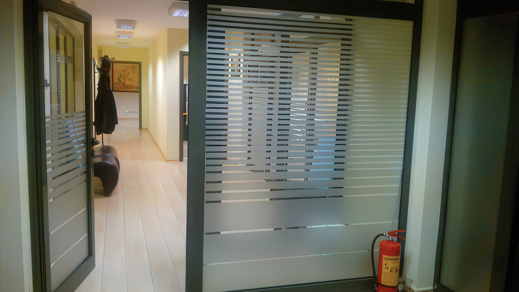 Офис интериор с матирани стъклени панели, отворена врата, закачалка за дрехи и пожарогасител до стената.