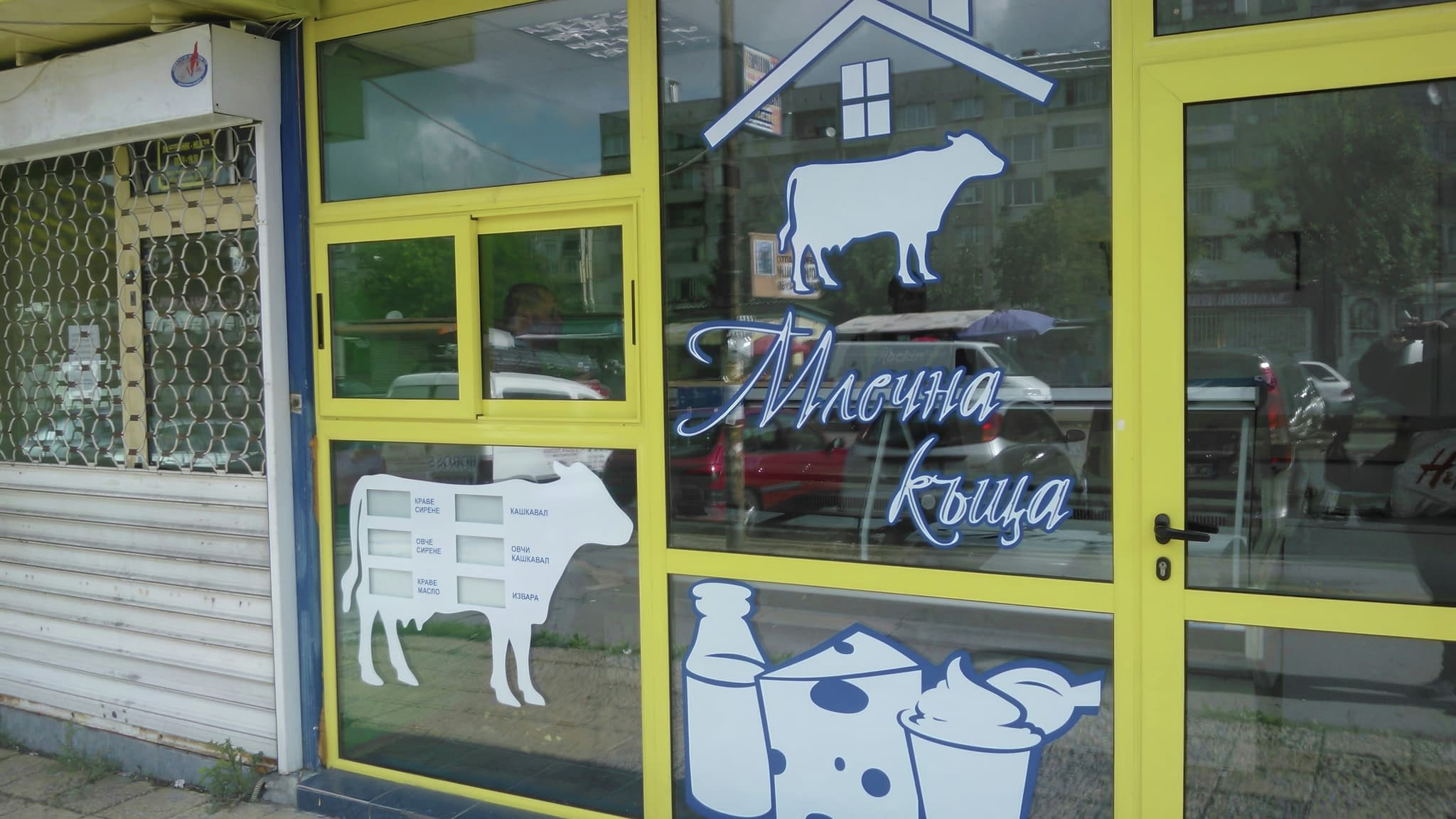 Витрина на магазин с илюстрация на крава и млечни продукти, с жълти рамки и сини акценти.