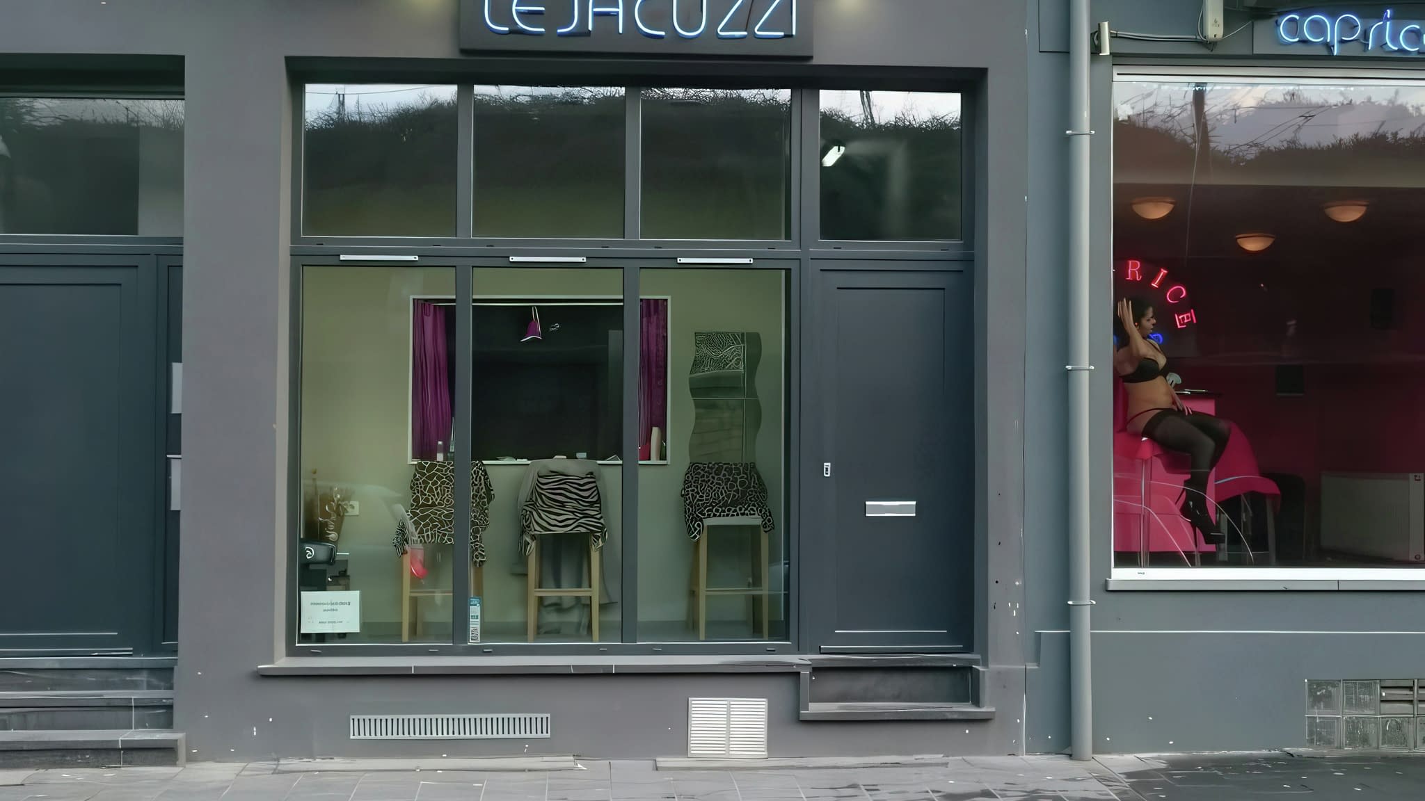 Дневен изглед към фасадата на „Le Jacuzzi“ с неонова табела и витрини, показващи зебра-декорирани столове, до съседен магазин с модни манекени.