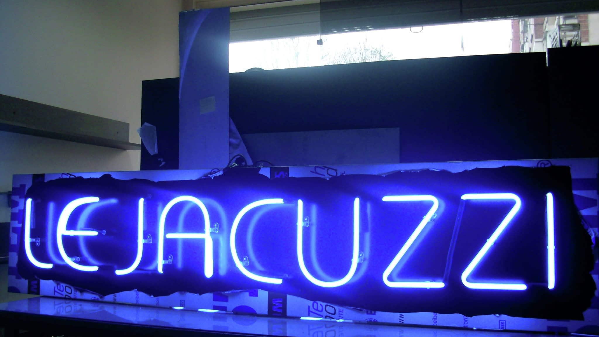 Ярка синя неонова табела с надпис „LE JACUZZI“, излъчваща светлина върху черен фон – пример за модерна неонова реклама.