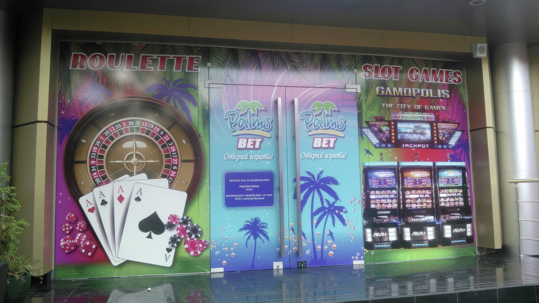 Вход на казино „Palms Bet“, облепен с рекламно фолио с изображения на рулетка, покер карти и слот машини.