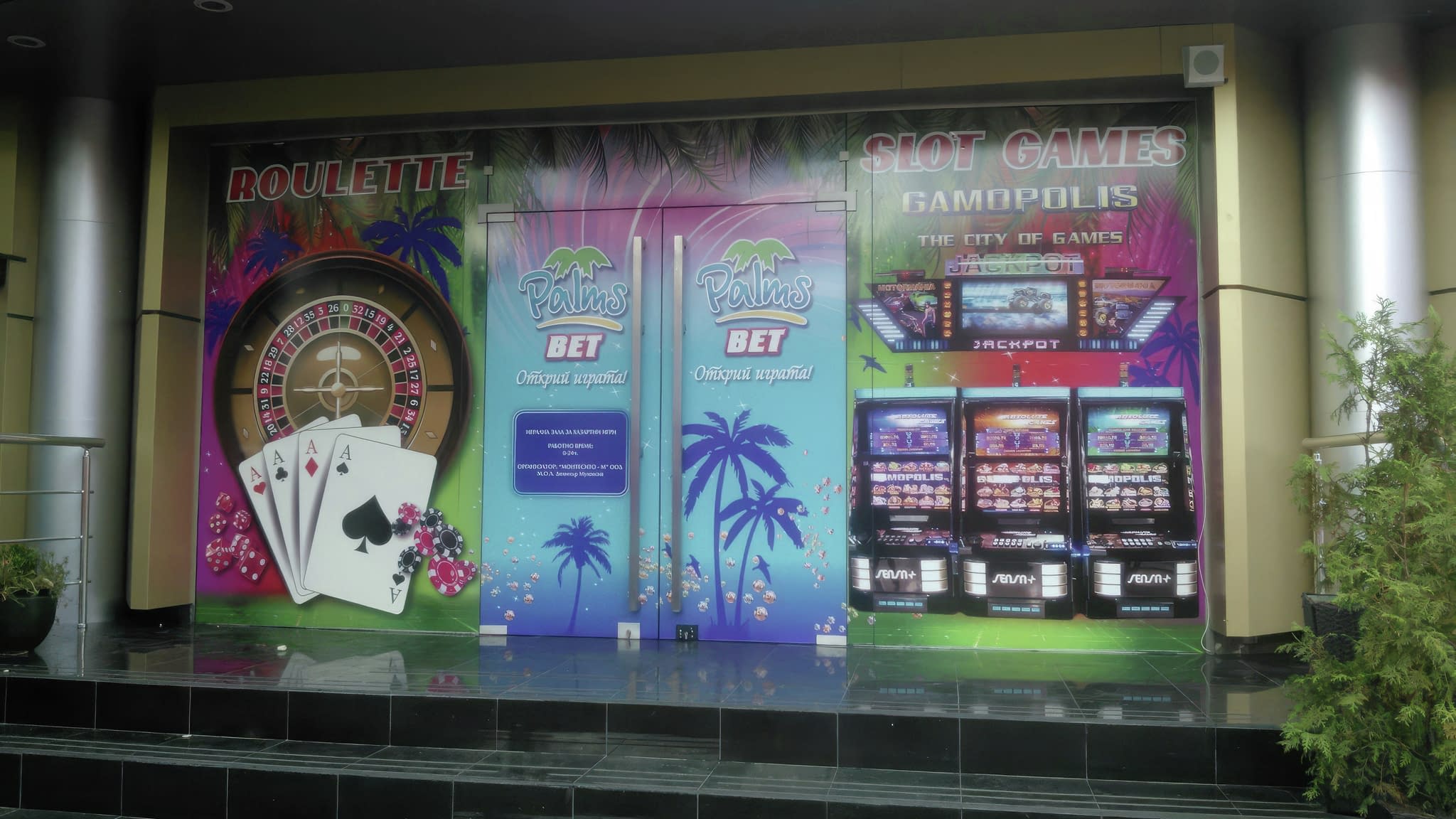 Облепен с рекламно фолио вход на казино с графики на рулетка, слот машини и реклама на „Palms Bet“.