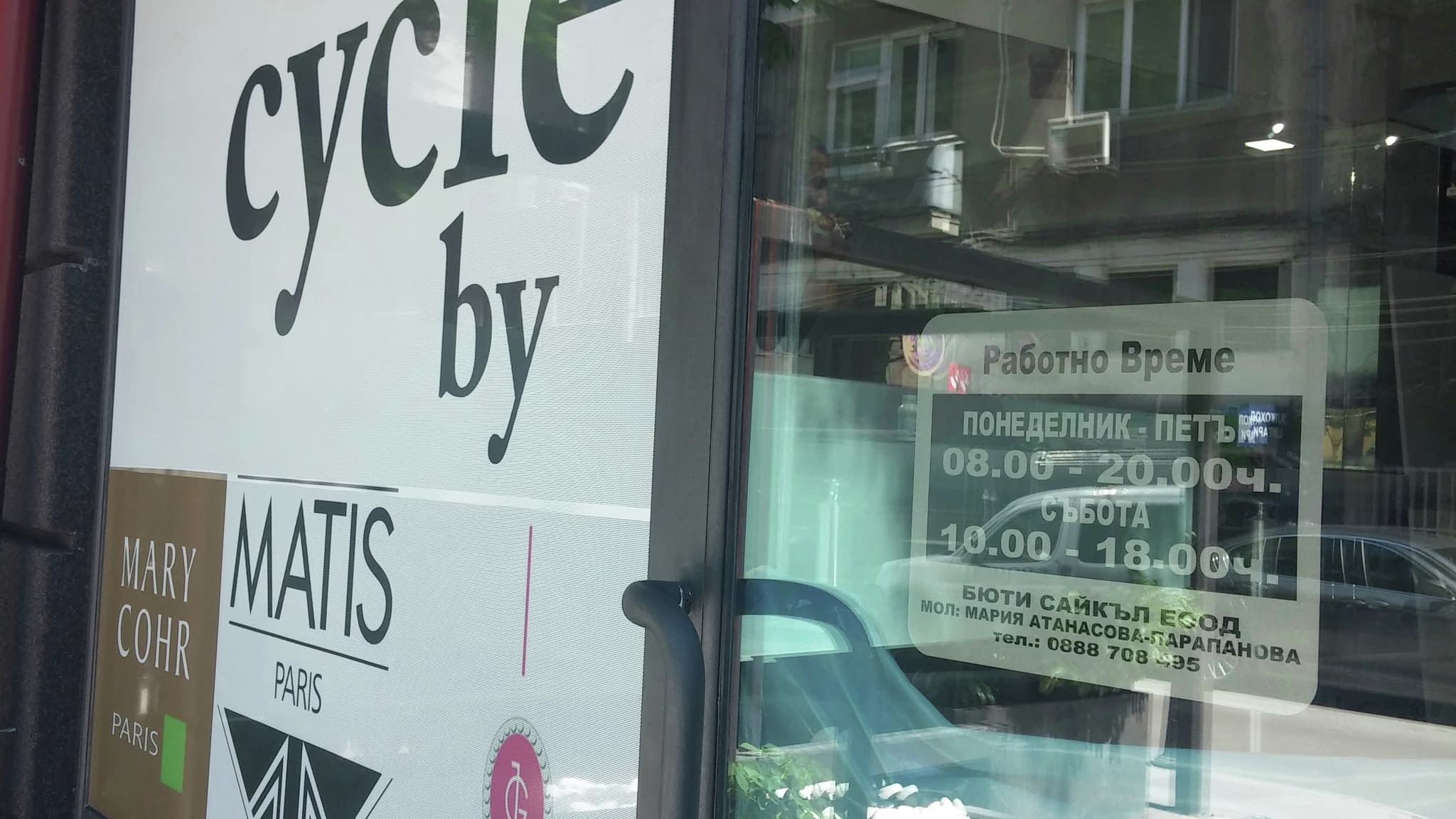 Витрина с перфо фолио, представяща бранда „Beauty Cycle“ и работно време на салона, заснета от ъгъл с отразени сгради във фона.