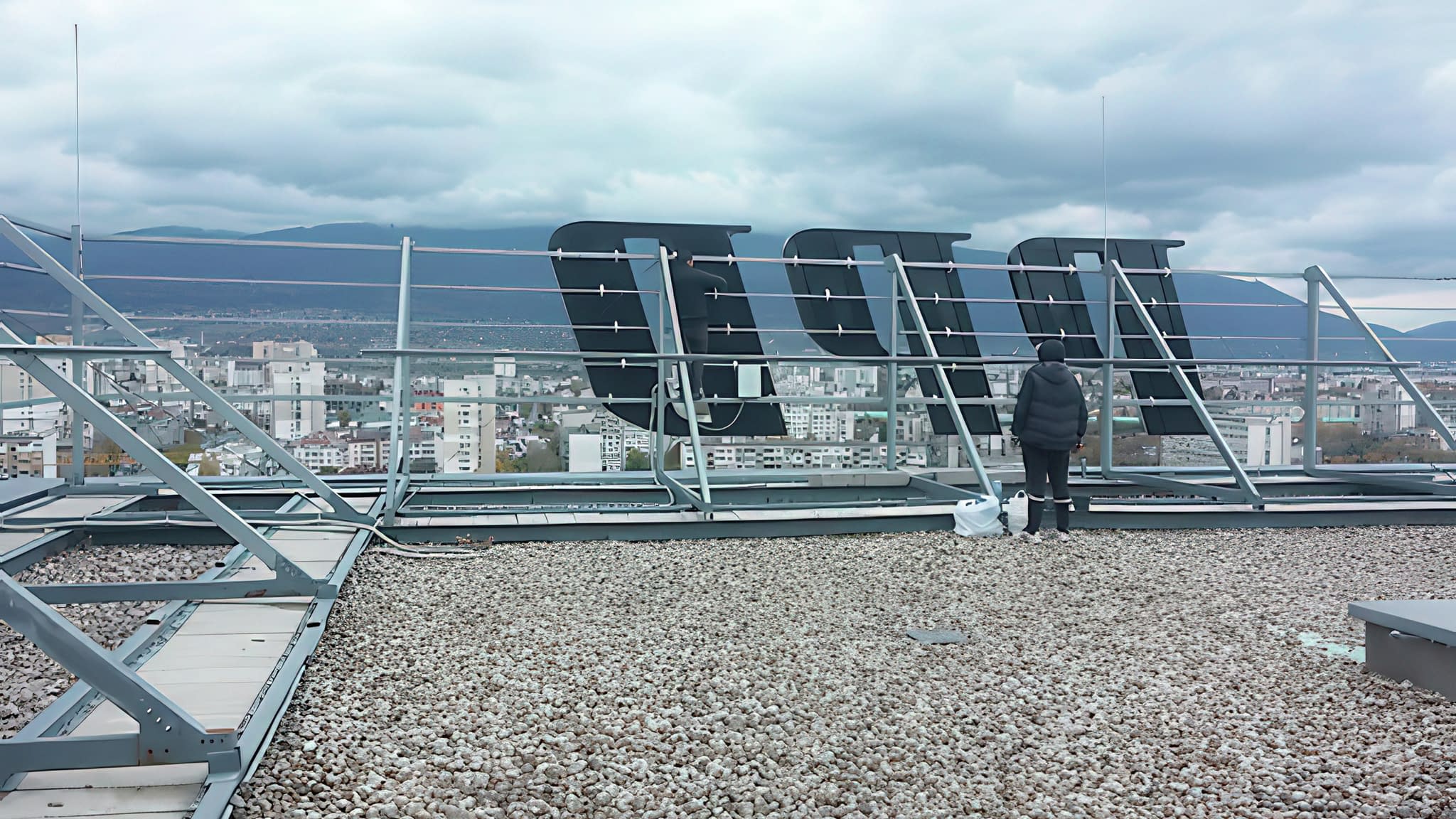 Монтаж на големи светещи букви върху покрива на висока сграда – работник инсталира LED надпис с панорамен изглед към града.