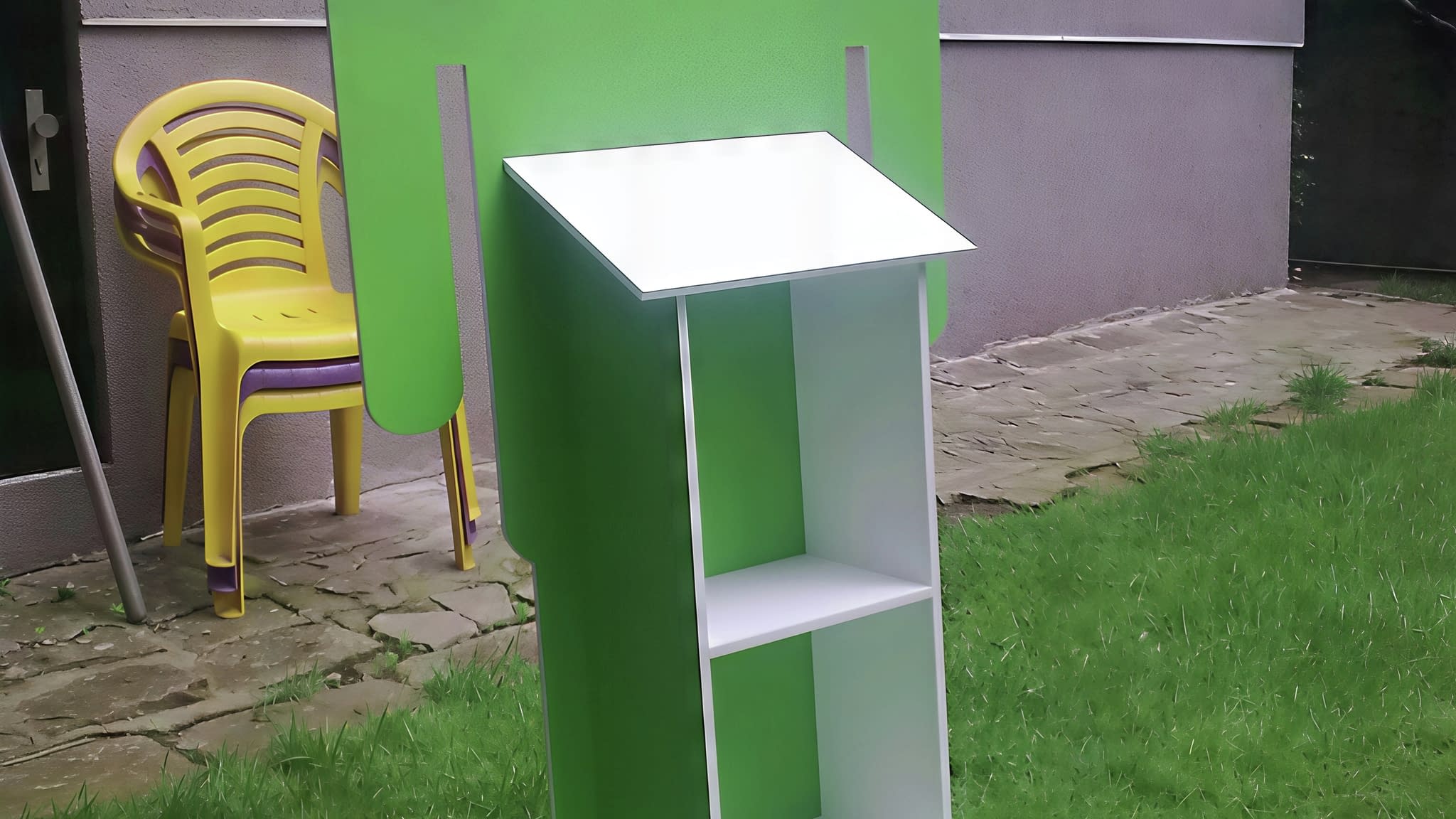 Голям рекламен стелаж със зелен Android талисман, бял рафт с три нива, разположен на тревна площ с жълт пластмасов стол на заден план.