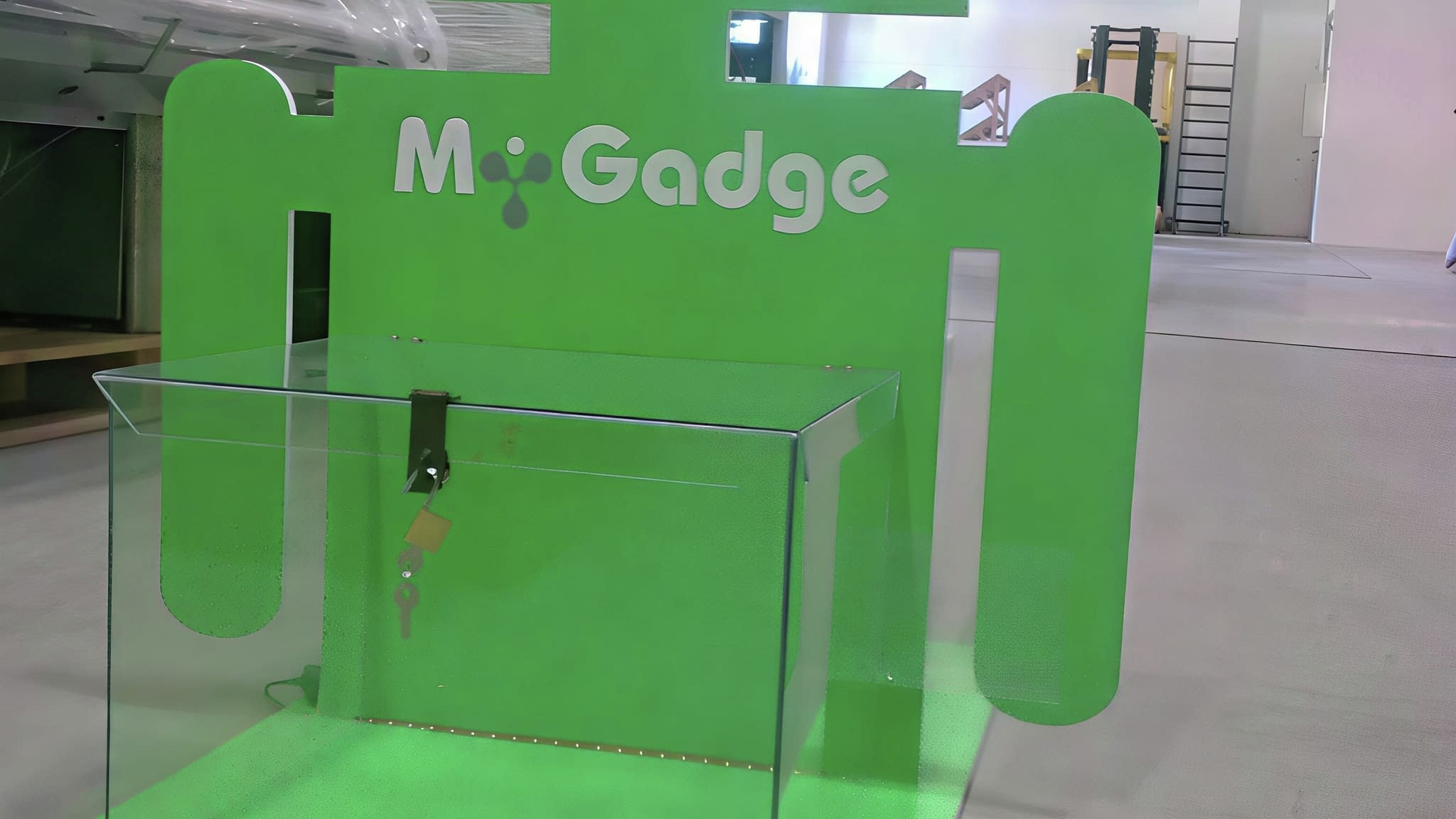Рекламен стелаж с форма на зелен Android талисман, с прозрачен шкаф с ключ за продукти и лого „M-Gadge“, поставен в индустриално хале.