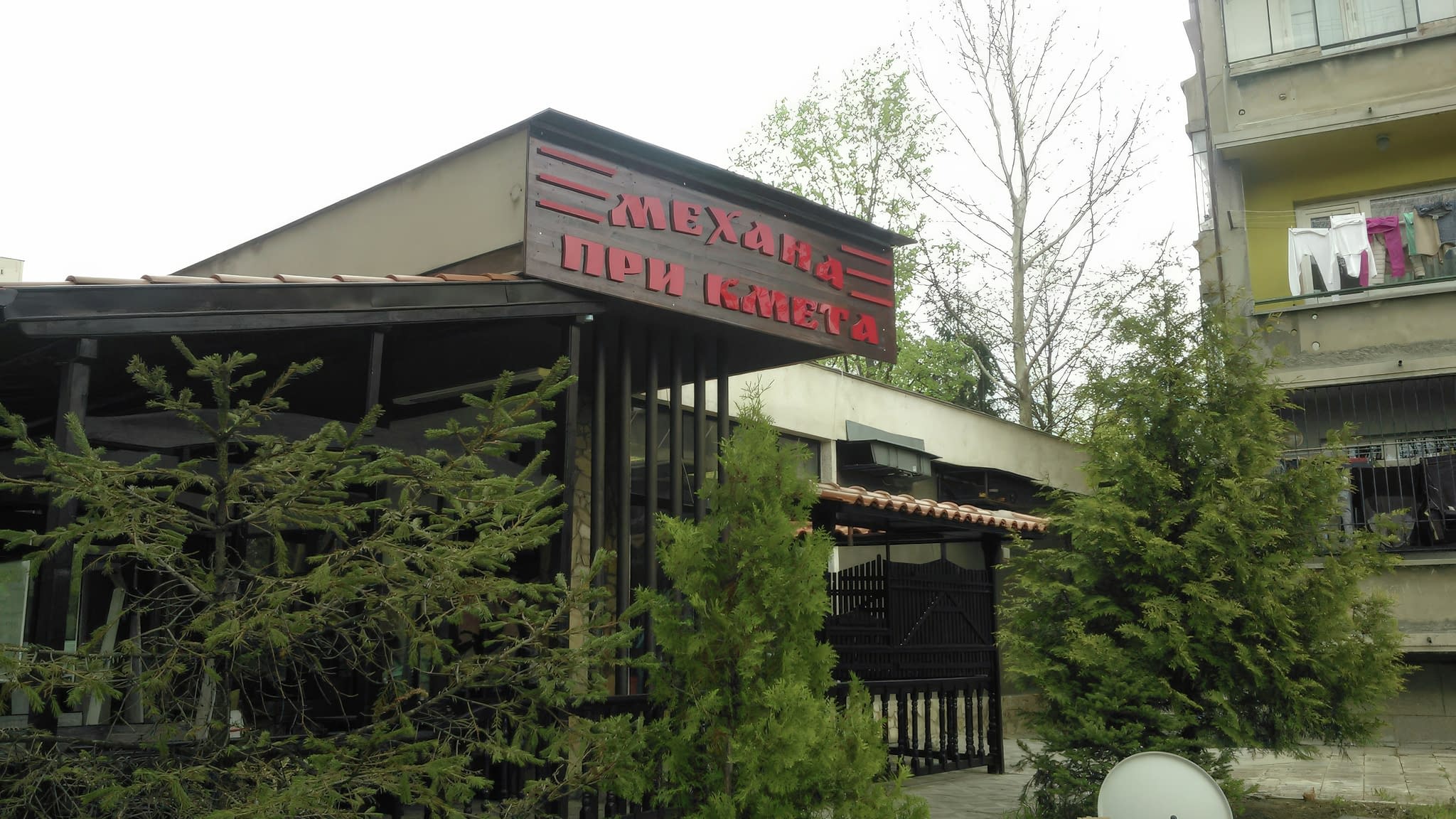 Фасада на ресторант „Механа при кмета“ с червена релефна табела и традиционна дървена архитектура сред зеленина.