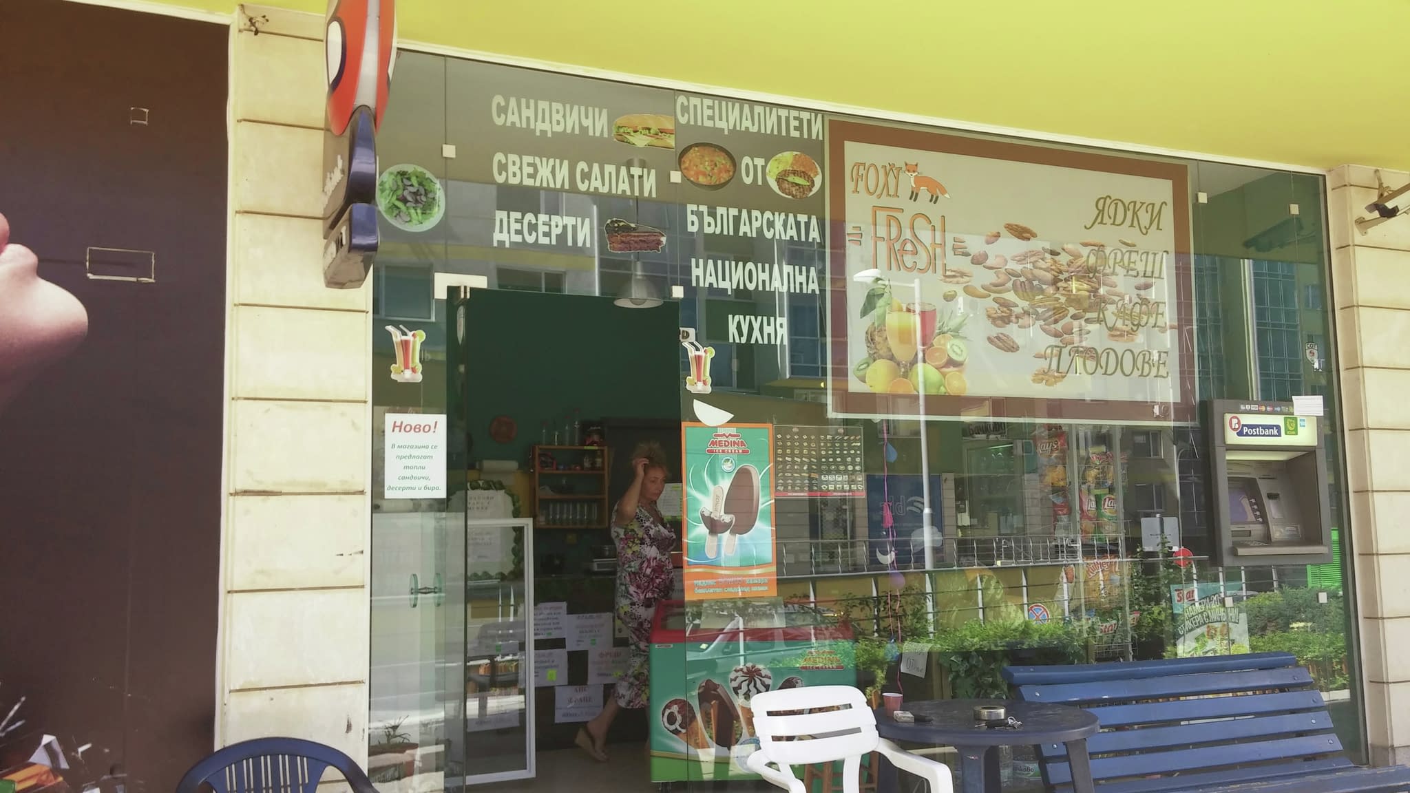 Външен изглед на кафене с рекламни стикери по стъклото, представящи меню със сандвичи, свежи салати и десерти, както и външна зона за сядане.
