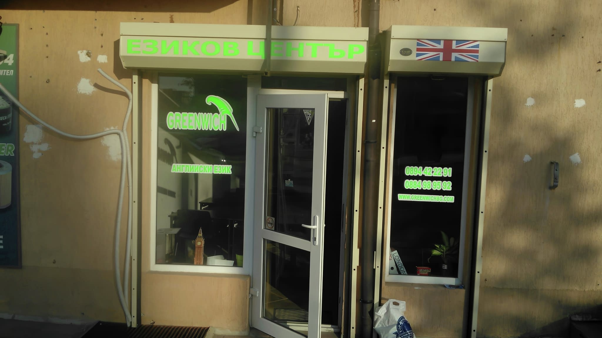 Рекламно фолио на витрина на езиков център „Greenwich“ със зелени надписи и британско знаме.
