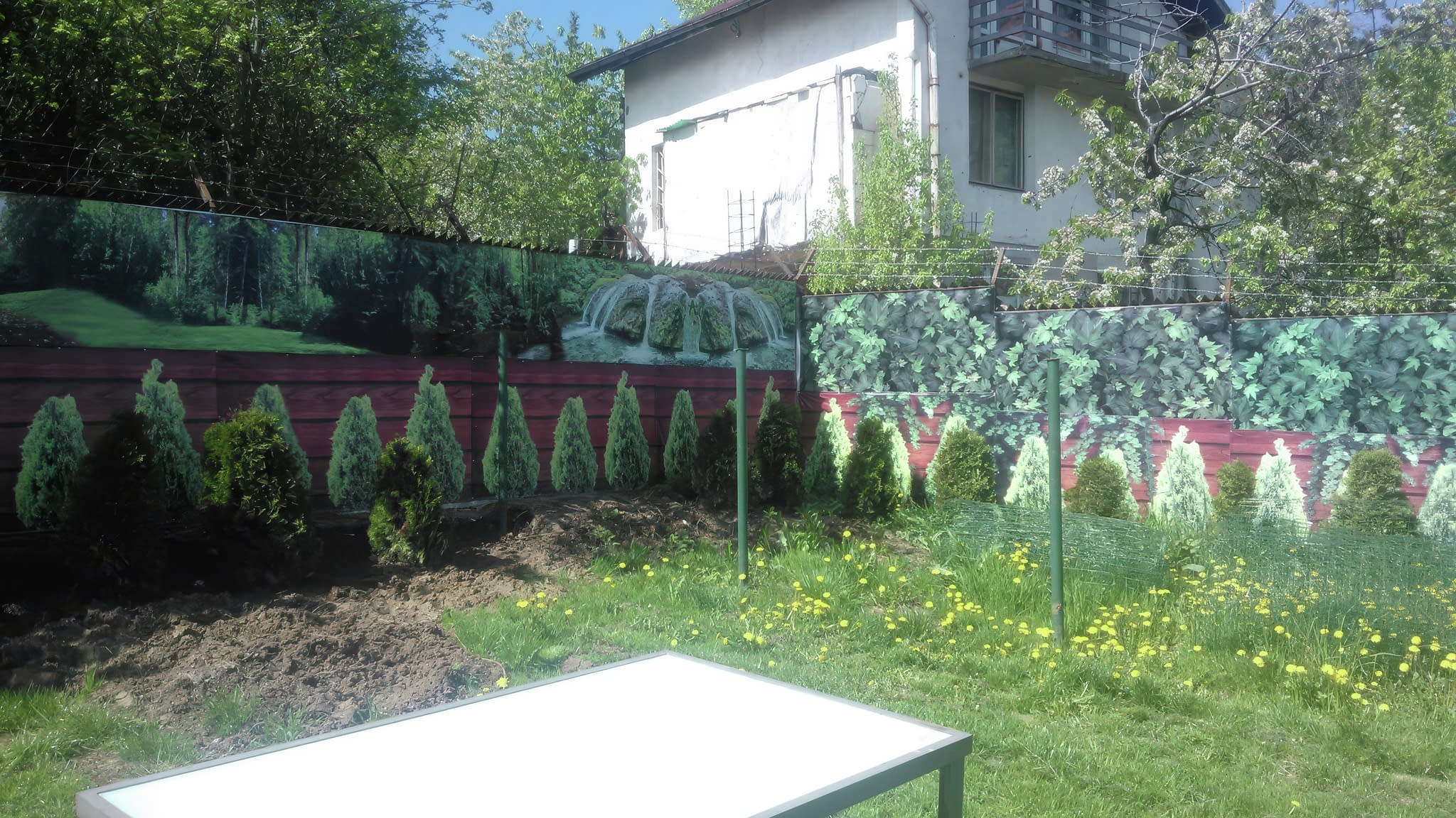 Винилно рекламно пано с природен пейзаж, монтирано на ограда в озеленен двор с декоративни храсти.