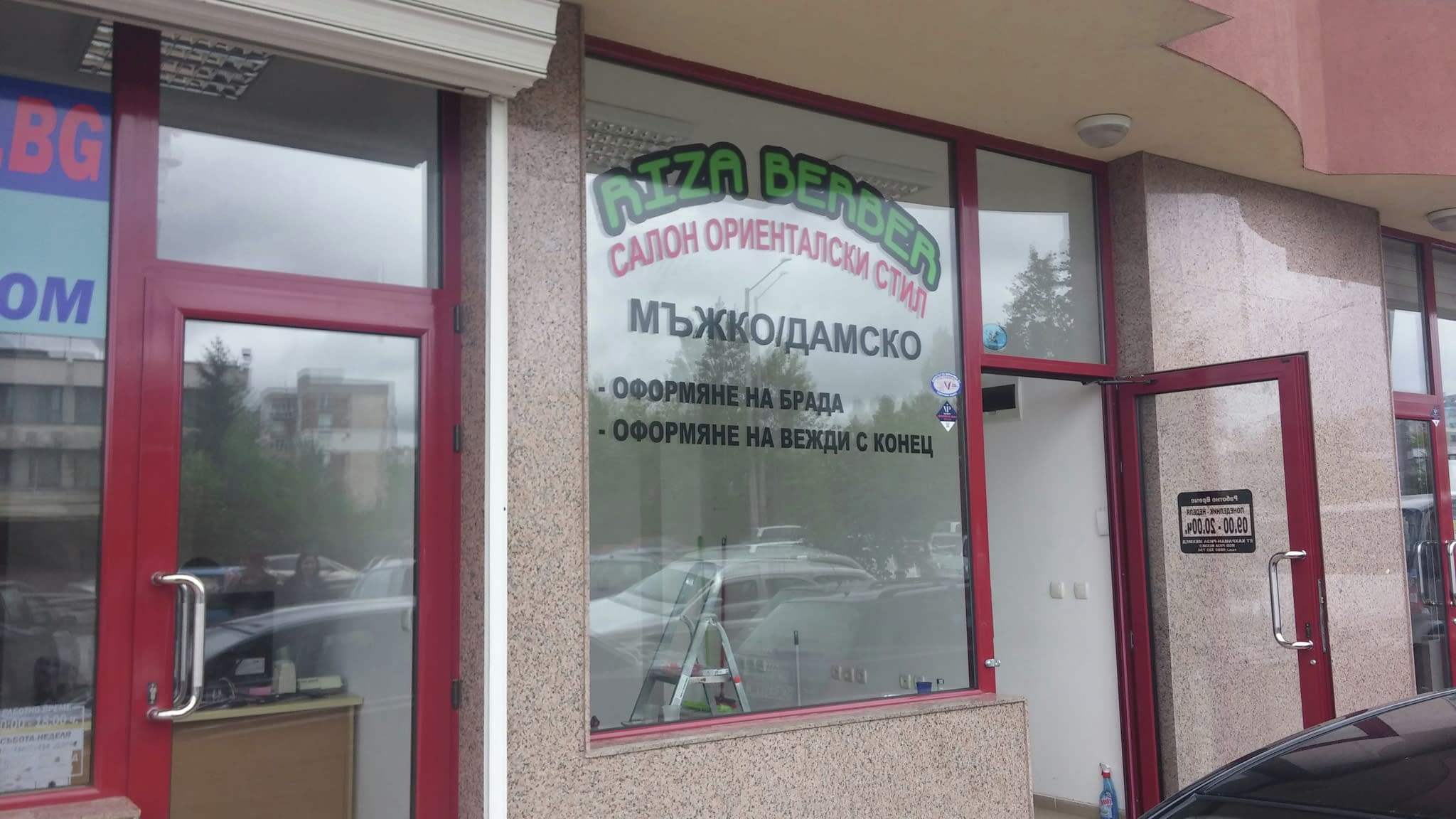 Външен изглед на бръснарница с рекламни стикери на витрината, представящи услуги като оформяне на брада и вежди, мъжки и дамски прически.