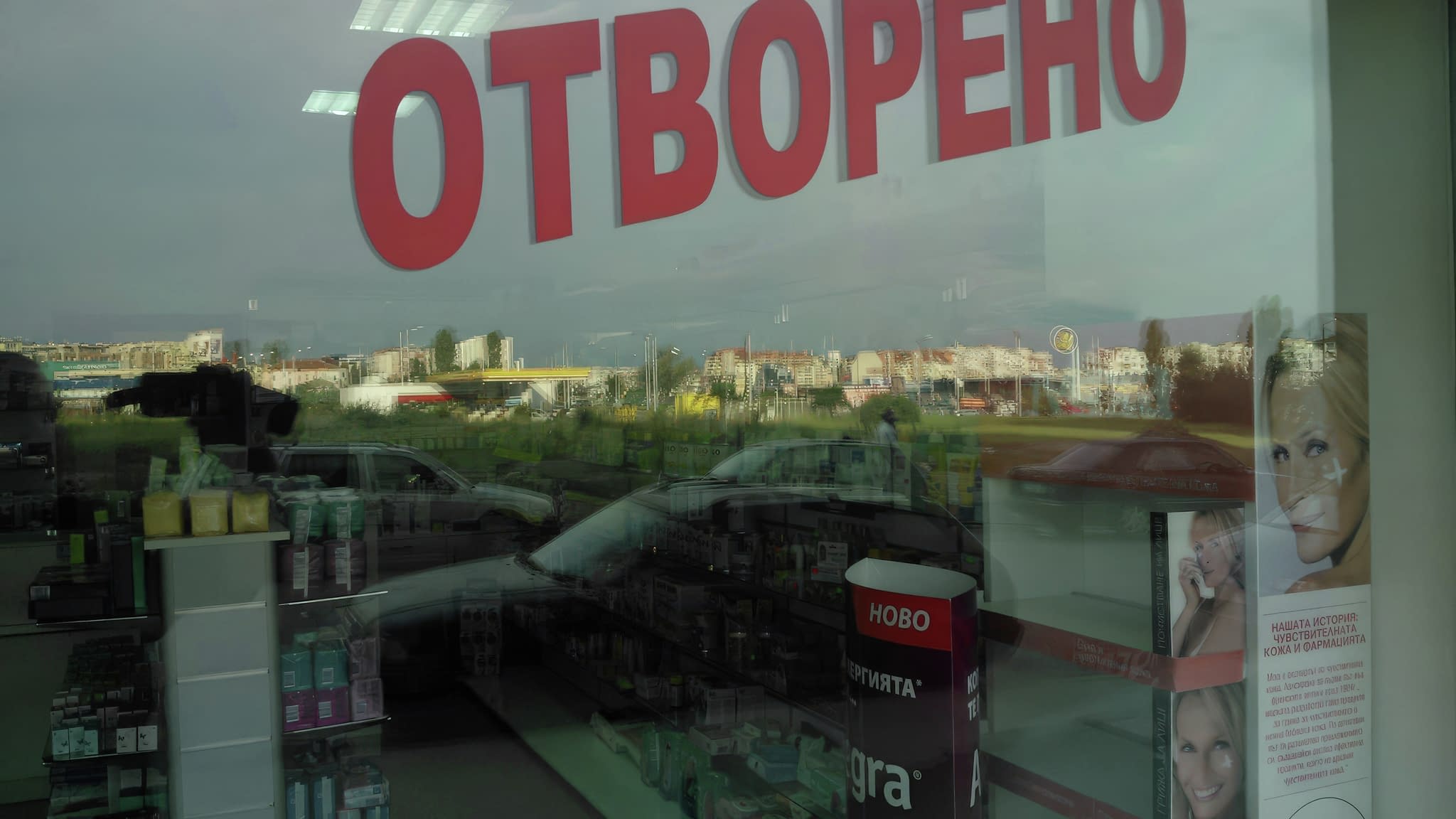 Стъклена витрина с рекламна лепенка с надпис ‘Отворено’ в големи червени букви, отразяваща градска среда и вътрешността на магазина с козметични продукти.