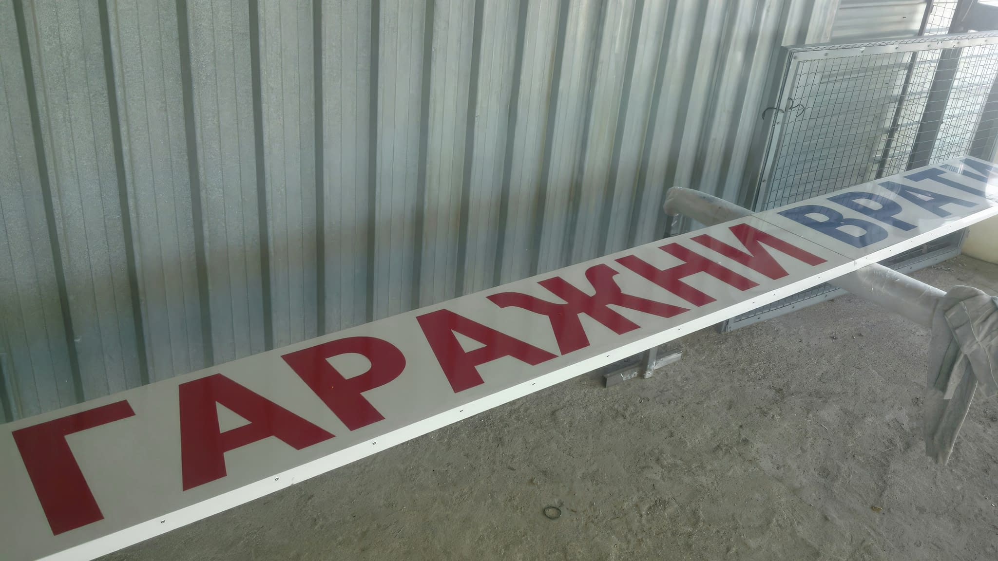 Рекламна табела за гаражни врати – бяла метална табела с надпис „Гаражни врати“ в червени и сини букви, поставена на стойки в работилница преди монтаж.