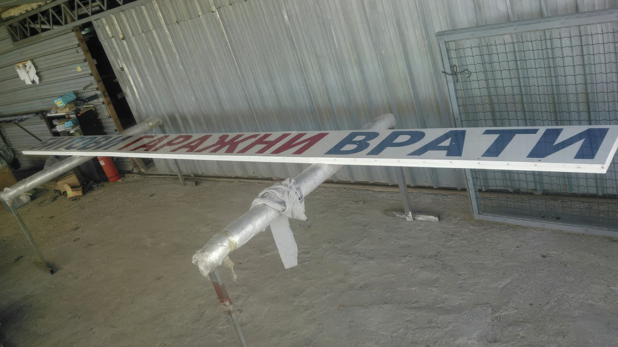 Изработка на информационна табела за гаражни врати – дълга метална табела с надпис „Гаражни врати“ в червени и сини букви, подготвена за монтаж в индустриално хале.