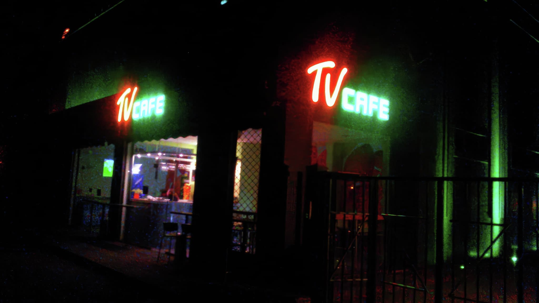 Нощен изглед към TV Cafe с обемни неонови букви в оранжево и зелено, осветяващи входа.
