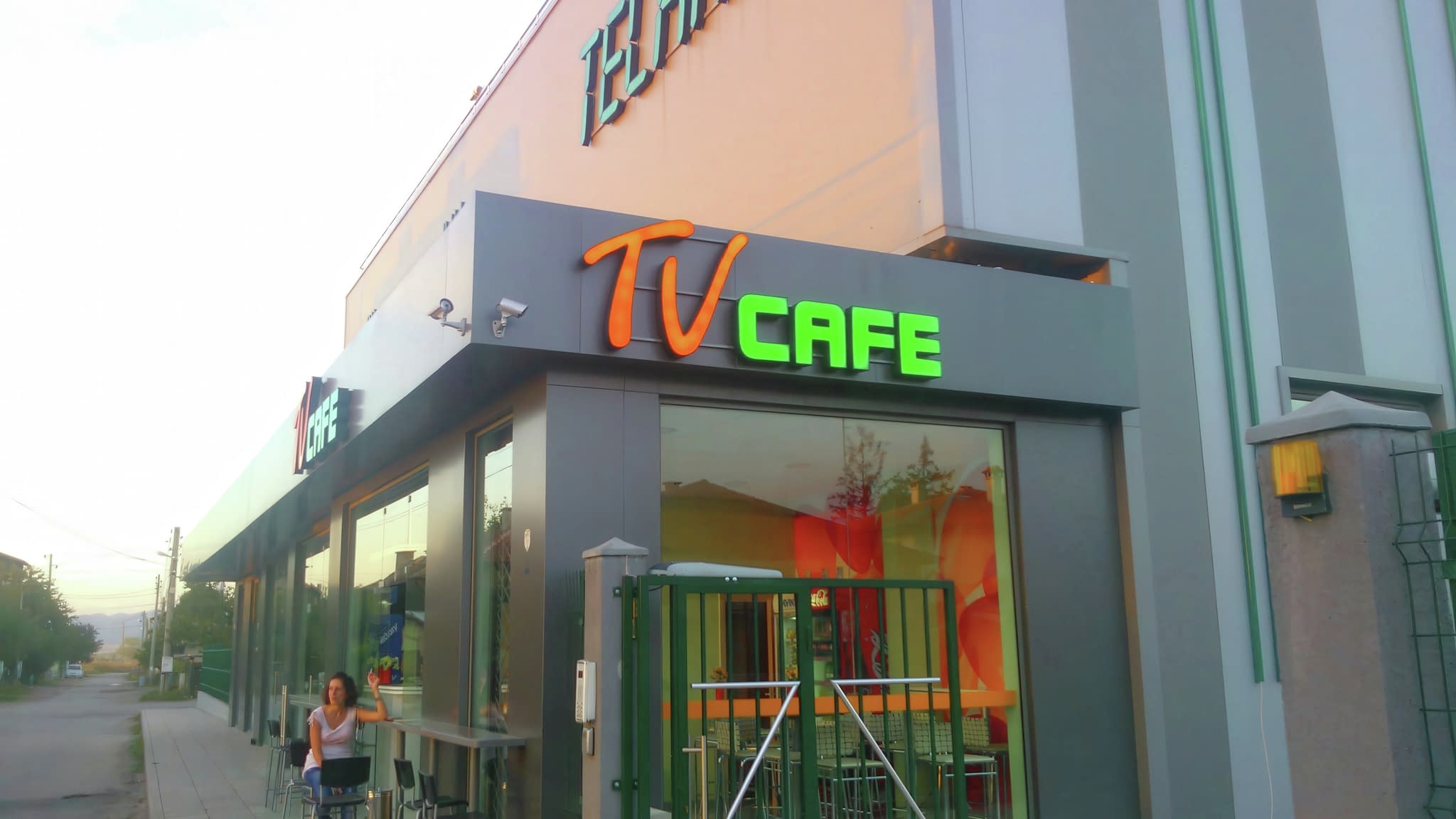 Фасада на TV Cafe с обемни светещи букви в оранжево и зелено, показващи логото, заснета през деня.