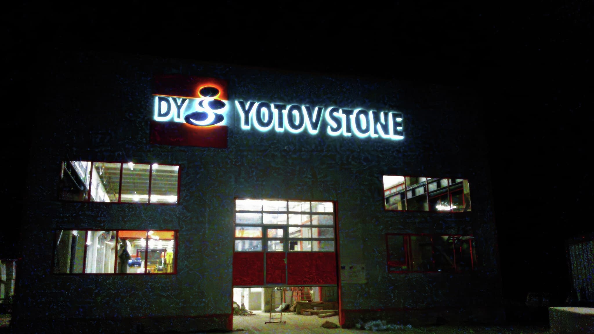Светещо лого „Yotov Stone” върху сграда през нощта.