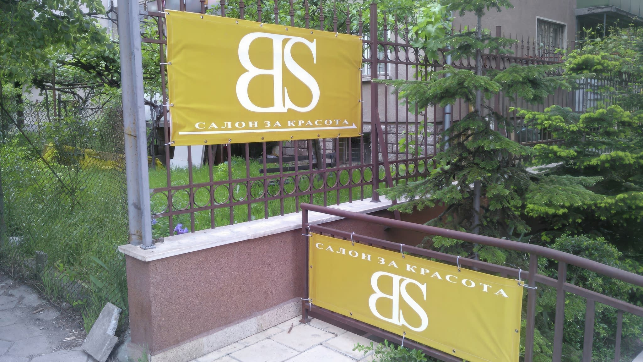 Две винилови рекламни пана с надпис 'Салон за красота' и логото 'BS', поставени на метална ограда пред входа на салон.