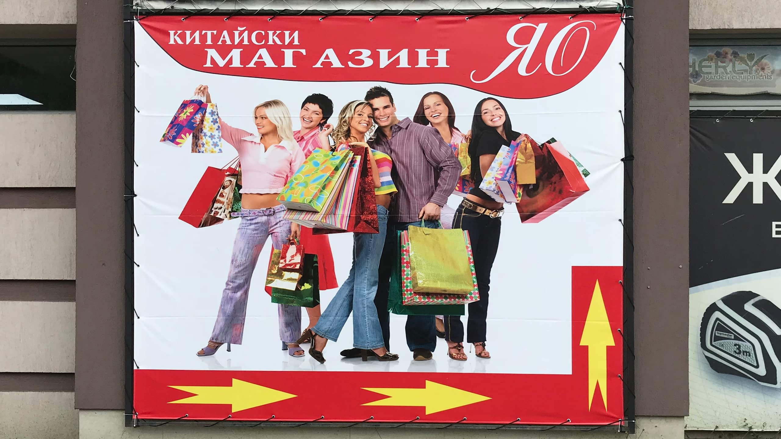 Рекламно платно за "Китайски магазин ЯО" с изображение на пазаруващи хора. Банерът служи и като указателна табела с жълти стрелки.