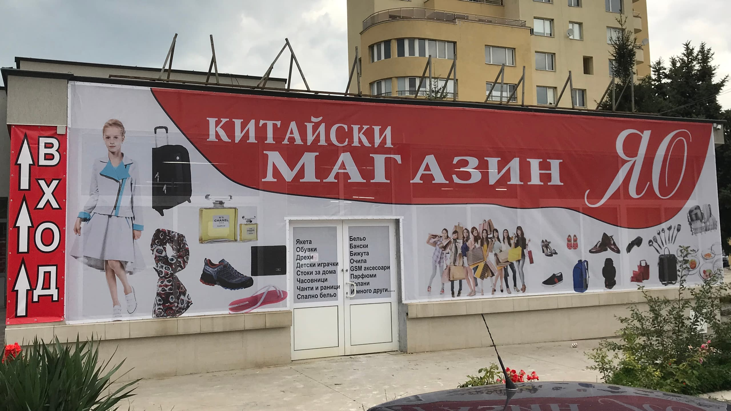 Голямо рекламно платно от винил за "Китайски магазин ЯО", опънато на фасадата на сграда. Банерът е червен и бял и показва различни видове стоки.