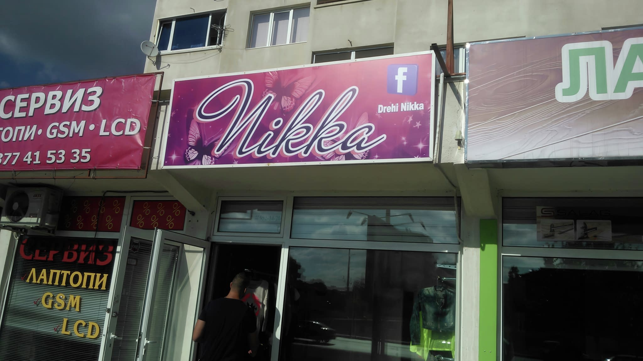 Винилова реклама с надпис 'Nikka' на лилав фон с пеперуди, монтирана над магазин за дрехи.